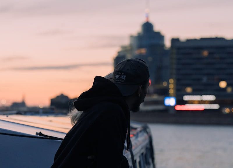 Man in a hoodie gazes over a city waterfront at sunset, blending urban tranquility.