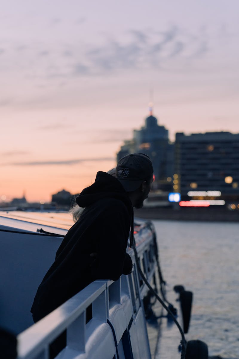 Man in a hoodie gazes over a city waterfront at sunset, blending urban tranquility.