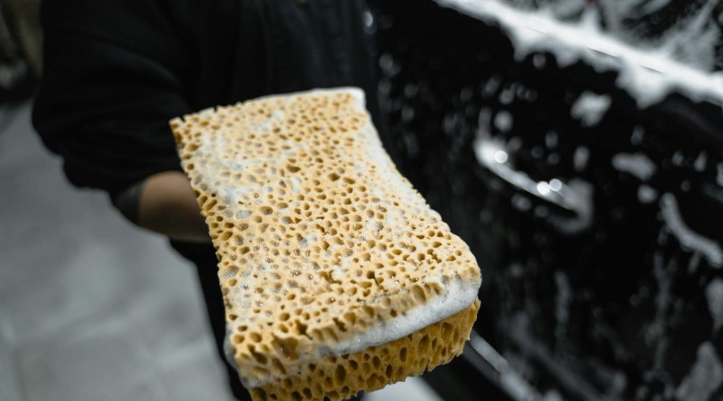 Sponge covered in soap suds held beside a car, highlighting car cleaning.